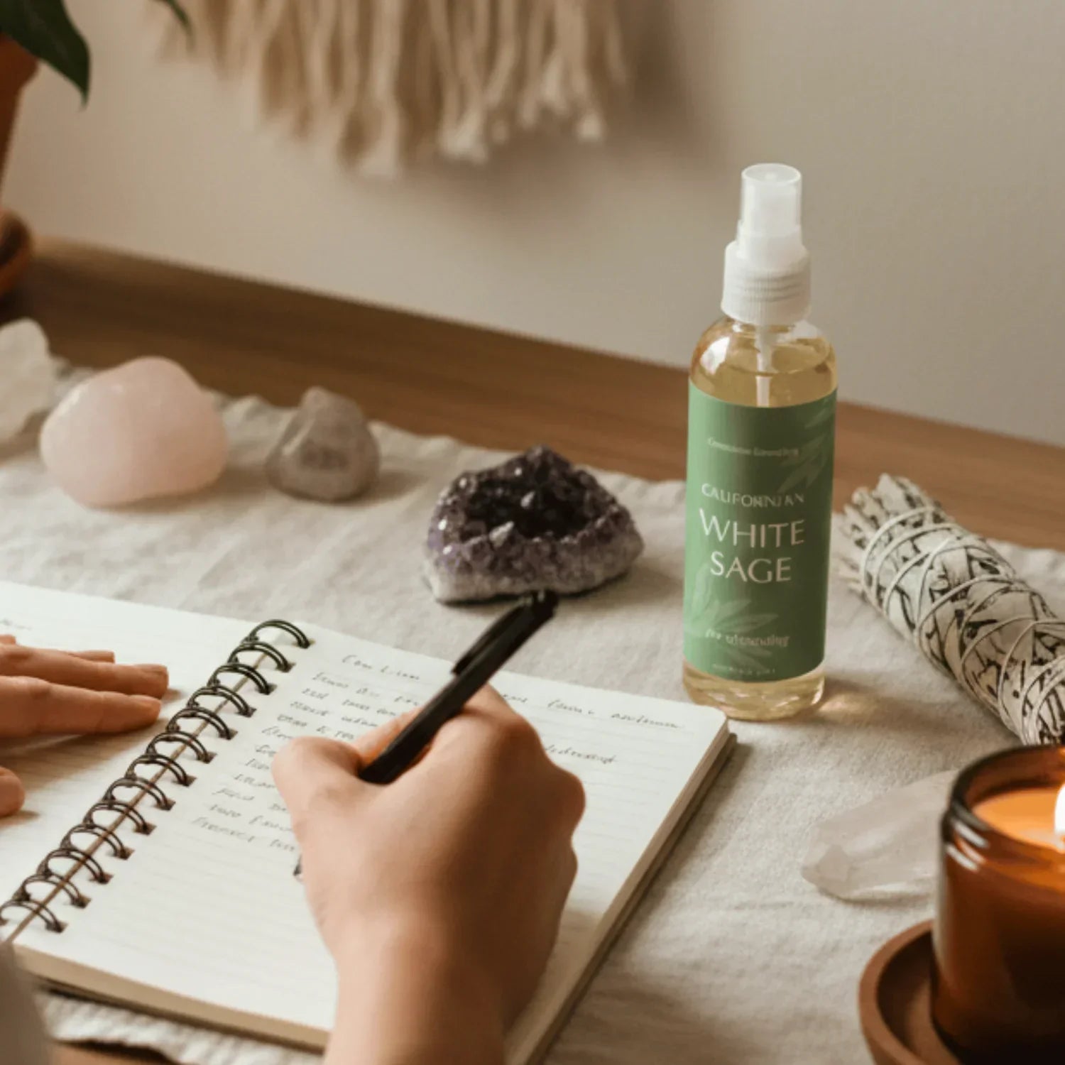 Persoon schrijft in journal naast witte salie spray, edelstenen en brandende kaars op tafel.